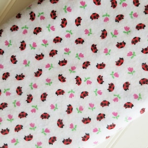 May include: White fabric with a repeating pattern of red ladybugs and pink and green flowers.