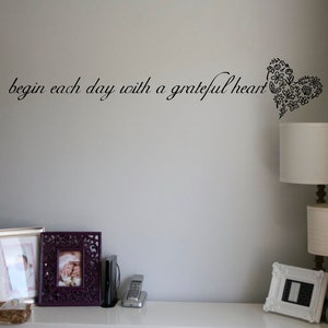 May include: A white wall with the quote "begin each day with a grateful heart" written in black cursive. A black floral heart design is to the right of the quote. There are three picture frames on a white shelf below the quote. The shelf is above a white dresser with drawers.