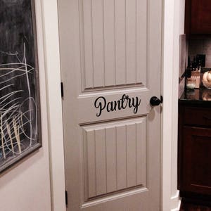 May include: A white door with a raised panel design and a black door handle. The door has a black vinyl sticker that says "Pantry" on it.