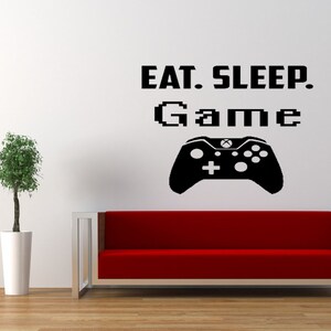 May include: A white wall with a black vinyl decal that reads "Eat. Sleep. Game." with a pixelated video game controller graphic. A red couch is in the foreground.