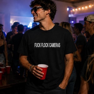 May include: A black short-sleeved t-shirt featuring the white text FUCK FLOCK CAMERAS printed across the chest. The shirt has a crew neckline and a standard fit, constructed from a solid black fabric with bold, high-contrast white lettering.
