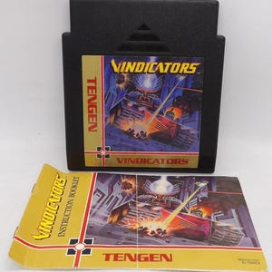 May include: A black Tengen Vindicators video game cartridge with a gold label featuring a futuristic tank illustration. The instruction booklet, also included, displays the game's title and artwork. The game is set against a futuristic backdrop.