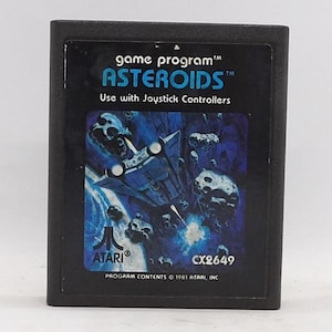 May include: A black Atari Asteroids game cartridge. The label features a blue spaceship graphic amid asteroids, with the game title and "Use with Joystick Controllers" text. The Atari logo and copyright information are also visible.