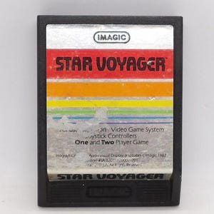 May include: A black Atari video game cartridge for "Star Voyager" by Imagic. The label features the game title in red, with a rainbow-colored stripe and the text "One and Two Player Game".