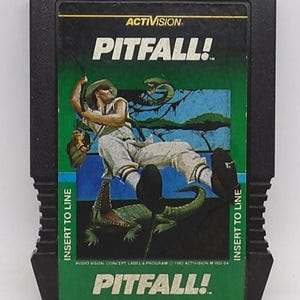 May include: Black Activision Pitfall! video game cartridge. The label features a man swinging on a vine, with a snake and crocodile. The game title is in large white letters. Green and blue accents.