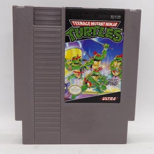 May include: A gray Nintendo Entertainment System (NES) cartridge for the game "Teenage Mutant Ninja Turtles." The label features the game's title and images of the four turtles in action, with a vibrant green and yellow color scheme.