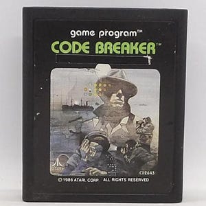 May include: Black Atari Code Breaker game cartridge. The label features a vintage illustration of a man in a hat, a ship, and other figures. The text "game program" and "CODE BREAKER" are displayed at the top.