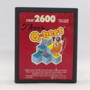 May include: A vintage Atari 2600 video game cartridge for Q*bert. The cartridge has a red label with the game's title in yellow and a cartoon character. The cartridge is black and has the text "D'Anna" written on it.