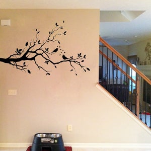 May include: Black silhouette of a tree branch with leaves and birds on a beige wall. The branch is on the left side of the image and extends towards the right. The wall is textured and has a light brown color.