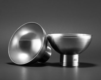May include: Two silver metal bowls with a rounded shape and a wide base. The bowl on the right has a small engraved logo on the base.