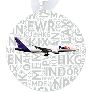 May include: A white round ornament with a FedEx airplane in flight. The ornament is covered in a pattern of airport codes, including DFW, HKG, KIX, MAD, MSP, ORD, and SJU.