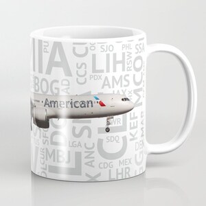 May include: White ceramic mug with a gray and white design featuring an American Airlines airplane and a list of airport codes. The airplane is flying over the text "American" with a red, white, and blue tail.