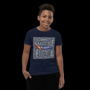 May include: Navy blue t-shirt featuring a Southwest airplane graphic. The design includes a collage of airport codes in white, such as DEN, LAS, and BWI. The shirt is worn by a person.