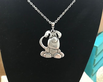 May include: Silver necklace with a charm of a cartoon dog sitting down. The dog has a big, round head with a small, black nose and big, round eyes.
