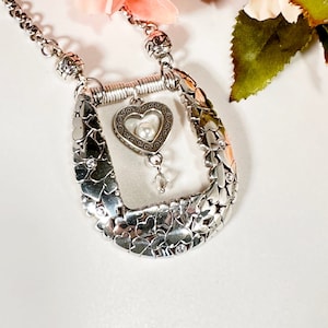 Pode incluir: Um colar de prata com um pingente em forma de cora&ccedil;&atilde;o que apresenta uma p&eacute;rola e um charme de cristal. O pingente &eacute; cercado por uma moldura de pequenos cora&ccedil;&otilde;es e pedras brilhantes. O colar est&aacute; sobre um fundo branco.