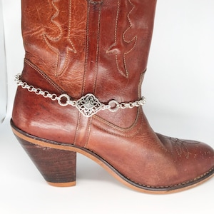 May include: Brown leather cowboy boot with a silver chain and decorative charm around the ankle.