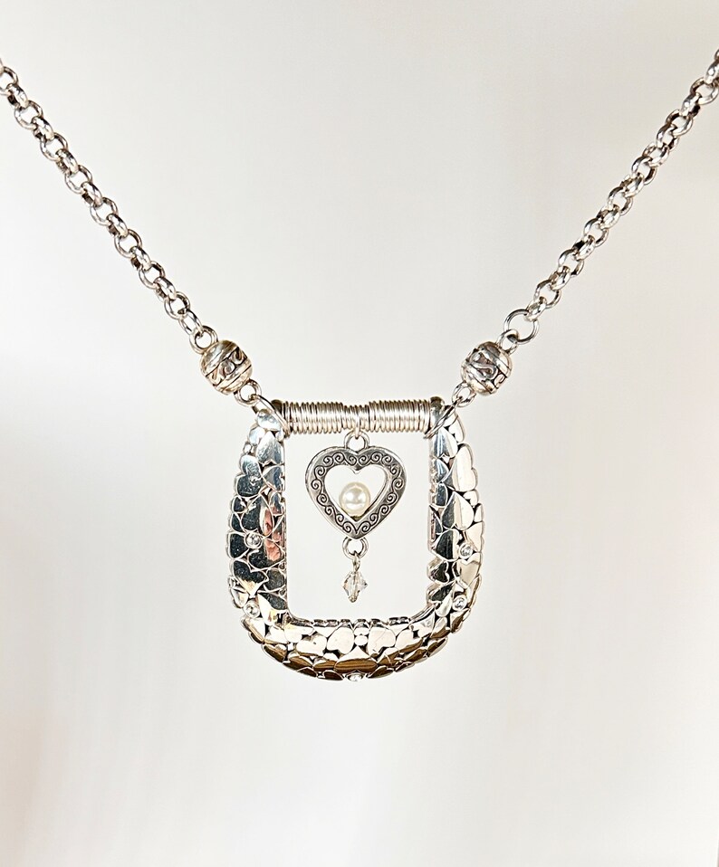 Pode incluir: Um colar de prata com um pingente em forma de cora&ccedil;&atilde;o. O pingente tem uma pequena p&eacute;rola no centro e &eacute; rodeado por um design delicado. O colar &eacute; feito de uma corrente com pequenas contas de prata.