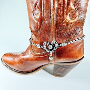 May include: Brown leather cowboy boot with a silver heart charm with rhinestones attached to a silver chain around the ankle.