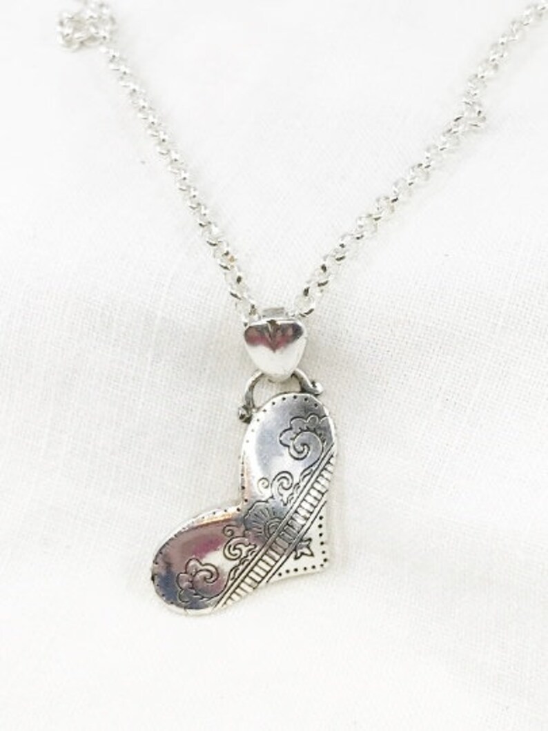 May include: A silver necklace with a heart-shaped pendant. The pendant has an intricate design of swirls and lines.