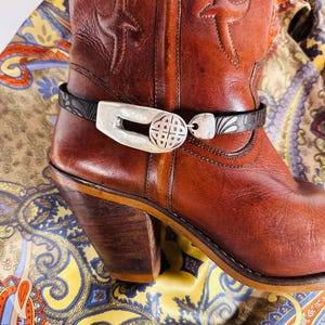 May include: A brown leather cowboy boot featuring a stacked wooden heel and decorative stitching. A black embossed leather strap wraps around the ankle, secured by a silver-toned metal buckle with a circular, intricate geometric cutout design.