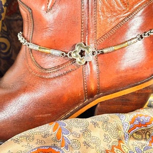 K&ouml;nnte beinhalten: Eine silber- und goldfarbene Stiefelkette mit einem bl&uuml;tenf&ouml;rmigen Anh&auml;nger ist &uuml;ber einem braunen Leder-Cowboystiefel drapiert. Der Stiefel liegt auf einem Schal mit Paisley-Muster und orangefarbenen, lila und gelben Akzenten.