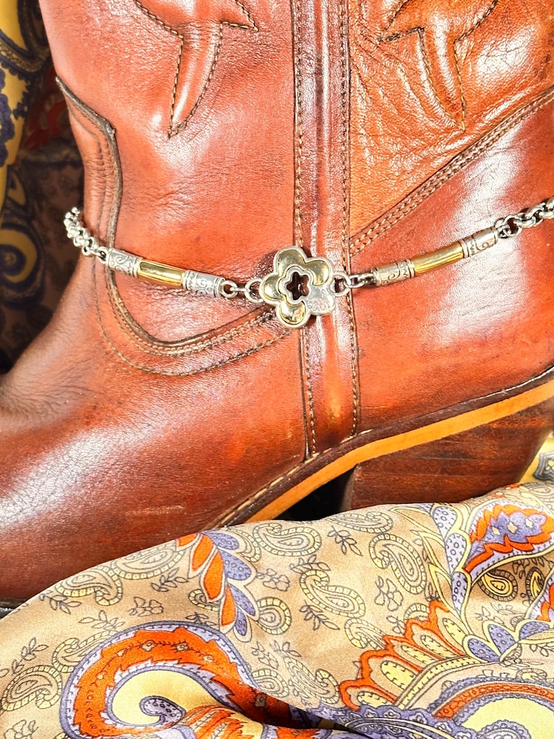 K&ouml;nnte beinhalten: Eine silber- und goldfarbene Stiefelkette mit einem bl&uuml;tenf&ouml;rmigen Anh&auml;nger ist &uuml;ber einem braunen Leder-Cowboystiefel drapiert. Der Stiefel liegt auf einem Schal mit Paisley-Muster und orangefarbenen, lila und gelben Akzenten.