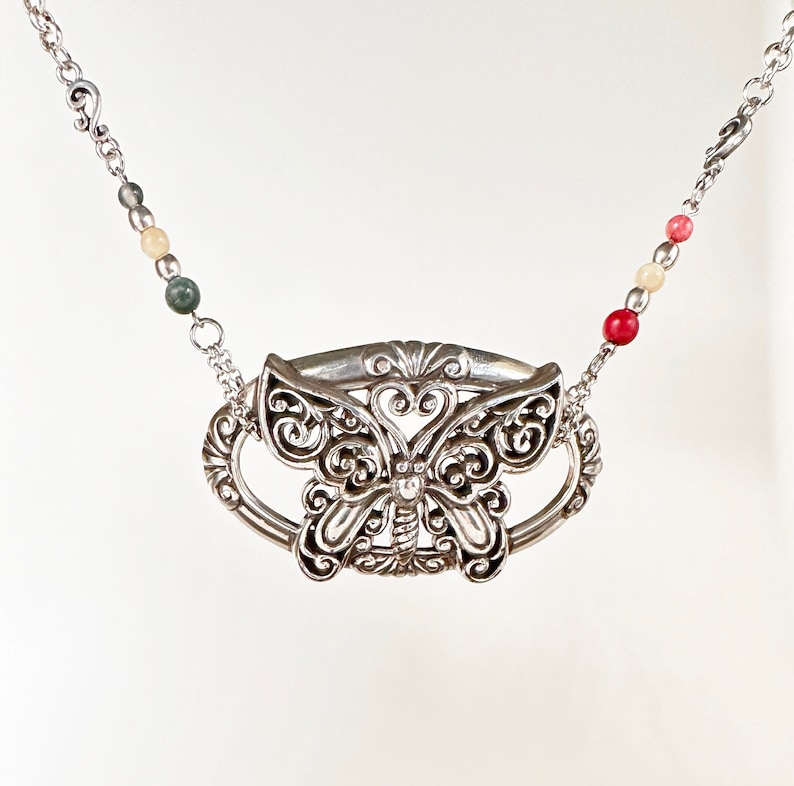 May include: A silver necklace with a butterfly pendant. The pendant is detailed with intricate filigree work and has a beaded chain with green, white, and red beads.