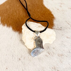 May include: A silver heart-shaped pendant with an intricate design hangs from a black cord necklace. The pendant is resting on a white rock.