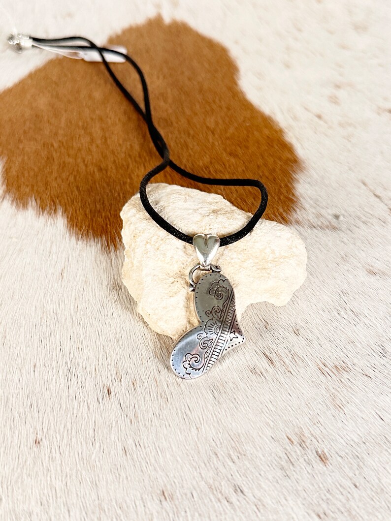 May include: A silver heart-shaped pendant with an intricate design hangs from a black cord necklace. The pendant is resting on a white rock.