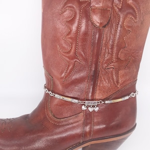 May include: A brown leather cowboy boot with a silver and gold ankle bracelet featuring a chain and heart charms.