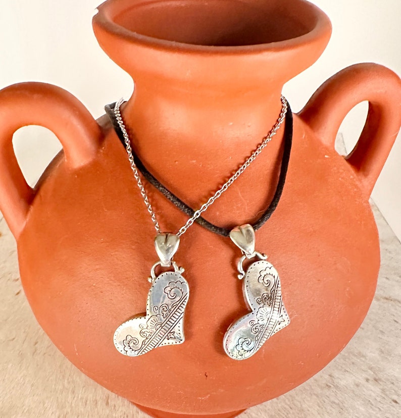 May include: Two silver heart-shaped pendants with intricate designs hang from a black cord and a silver chain. The pendants are hanging from a terracotta vase.