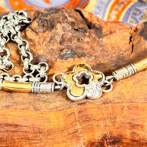 K&ouml;nnte beinhalten: Silber- und goldfarbene Halskette mit einem bl&uuml;tenf&ouml;rmigen Anh&auml;nger. Der Anh&auml;nger hat eine goldfarbene Mitte mit silbernem Umriss und einem kleinen Logo. Die Halskette liegt auf einer Holzoberfl&auml;che mit bunt gemustertem Stoff im Hintergrund.