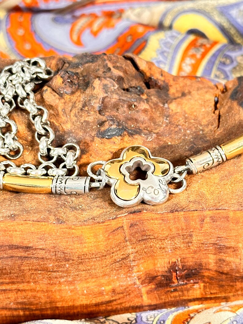 K&ouml;nnte beinhalten: Silber- und goldfarbene Halskette mit einem bl&uuml;tenf&ouml;rmigen Anh&auml;nger. Der Anh&auml;nger hat eine goldfarbene Mitte mit silbernem Umriss und einem kleinen Logo. Die Halskette liegt auf einer Holzoberfl&auml;che mit bunt gemustertem Stoff im Hintergrund.