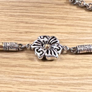 K&ouml;nnte beinhalten: Silberfarbene Halskette mit einem Blumenanh&auml;nger. Die Blume hat eine goldene Mitte und detaillierte silberne Bl&uuml;tenbl&auml;tter. Die Halskette hat goldene und silberne Akzente und einen Kettenverschluss. Der Schmuck wird auf einer hellen Holzoberfl&auml;che pr&auml;sentiert.