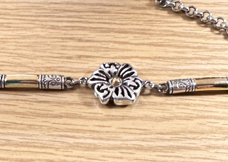 K&ouml;nnte beinhalten: Silberfarbene Halskette mit einem Blumenanh&auml;nger. Die Blume hat eine goldene Mitte und detaillierte silberne Bl&uuml;tenbl&auml;tter. Die Halskette hat goldene und silberne Akzente und einen Kettenverschluss. Der Schmuck wird auf einer hellen Holzoberfl&auml;che pr&auml;sentiert.