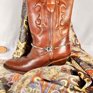 K&ouml;nnte beinhalten: Ein brauner Leder-Cowboystiefel mit einer silbernen und schwarzen Stiefelkette. Der Stiefel ist mit gestickten Mustern verziert. Ein farbenfrohes Paisley-Tuch ist um den Stiefel drapiert.