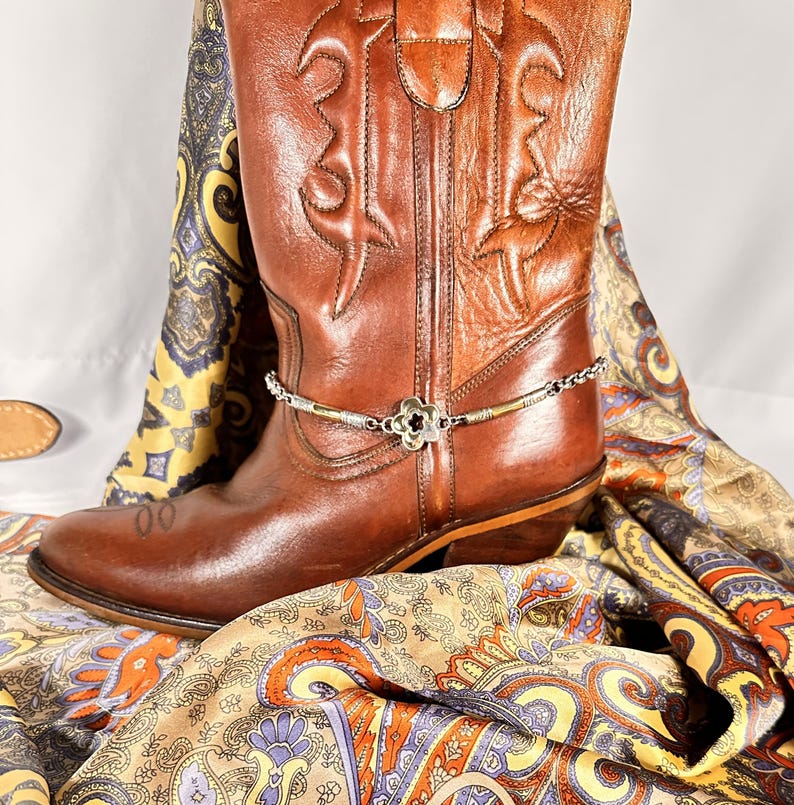 K&ouml;nnte beinhalten: Ein brauner Leder-Cowboystiefel mit einer silbernen und schwarzen Stiefelkette. Der Stiefel ist mit gestickten Mustern verziert. Ein farbenfrohes Paisley-Tuch ist um den Stiefel drapiert.