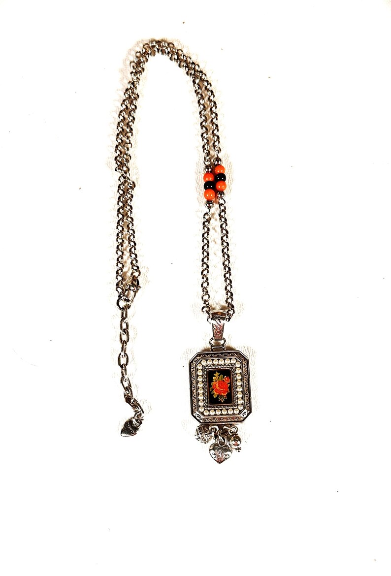 May include: Silver necklace with a rectangular pendant. The pendant has a red rose design on a black background, framed by faux pearls. The chain includes orange and black beads and heart charms.