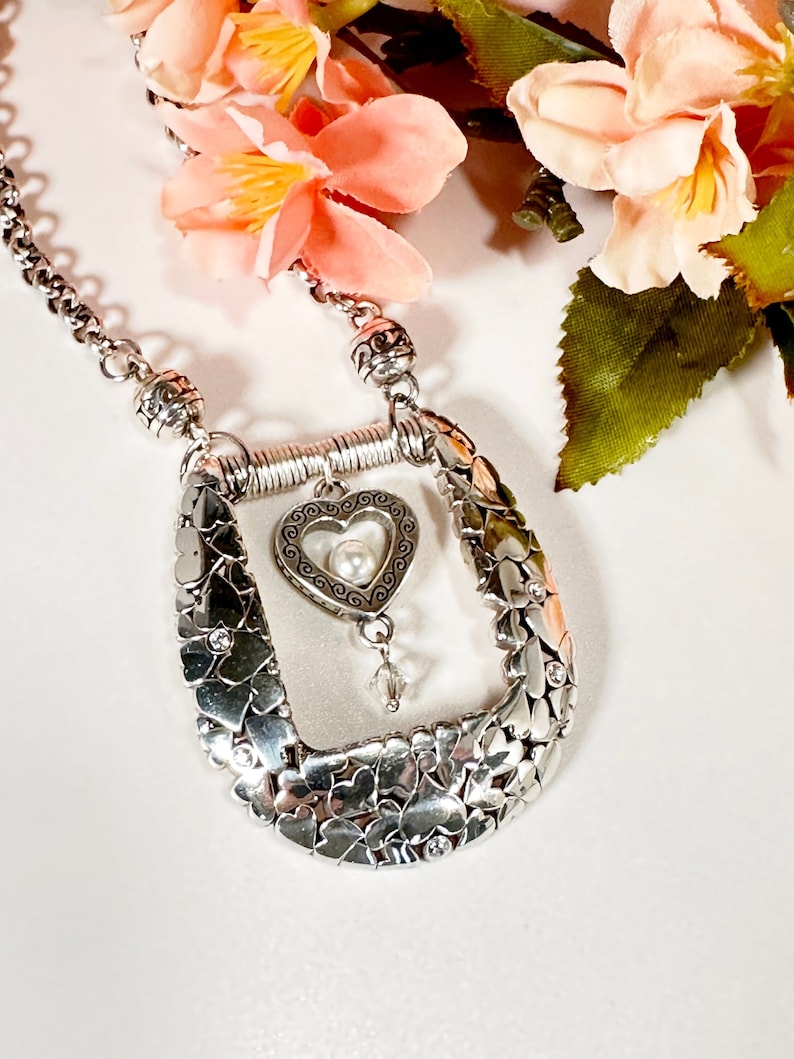 Pode incluir: Um colar de prata com um pingente em forma de cora&ccedil;&atilde;o que apresenta uma p&eacute;rola e um charme de cristal. O pingente &eacute; cercado por uma moldura de pequenos cora&ccedil;&otilde;es e pedras brilhantes. O colar est&aacute; sobre um fundo branco.