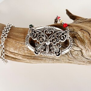 May include: A silver butterfly pendant necklace with a delicate filigree design. The necklace features a chain and a beaded strand with red, white, and cream colored beads.
