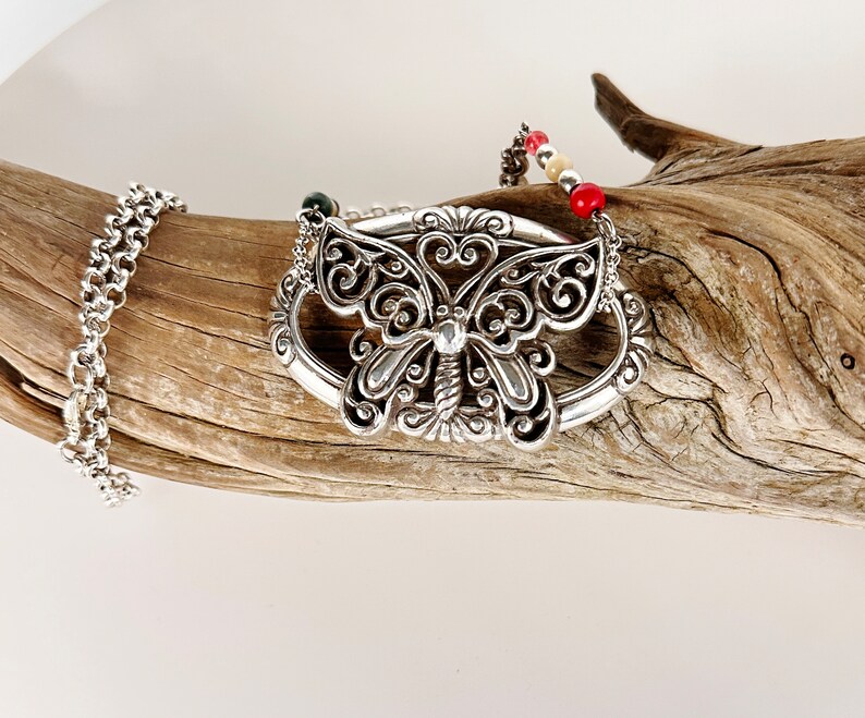 May include: A silver butterfly pendant necklace with a delicate filigree design. The necklace features a chain and a beaded strand with red, white, and cream colored beads.