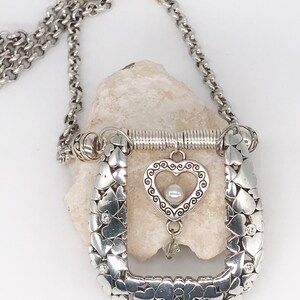 Pode incluir: Um colar de prata com um pingente em forma de cora&ccedil;&atilde;o. O pingente &eacute; feito de muitos cora&ccedil;&otilde;es pequenos e tem um cora&ccedil;&atilde;o maior no centro com uma p&eacute;rola. O colar est&aacute; em uma corrente de prata.