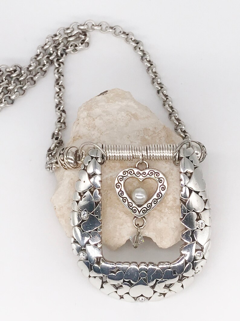 Pode incluir: Um colar de prata com um pingente em forma de cora&ccedil;&atilde;o. O pingente &eacute; feito de muitos cora&ccedil;&otilde;es pequenos e tem um cora&ccedil;&atilde;o maior no centro com uma p&eacute;rola. O colar est&aacute; em uma corrente de prata.