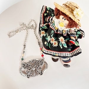 May include: A silver butterfly pendant necklace with a chain and red, green, and white beads. The necklace is on a white surface with a doll wearing a black and white dress and a straw hat.