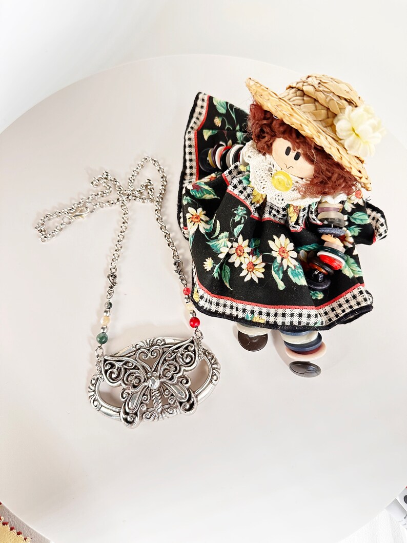 May include: A silver butterfly pendant necklace with a chain and red, green, and white beads. The necklace is on a white surface with a doll wearing a black and white dress and a straw hat.