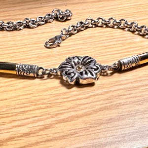 K&ouml;nnte beinhalten: Silberfarbenes Armband mit bl&uuml;tenf&ouml;rmigem Mittelst&uuml;ck und goldfarbenen Akzenten. Das Armband hat ein Kettenglied-Design und einen Karabinerverschluss. Die Blume hat eine goldfarbene Mitte.