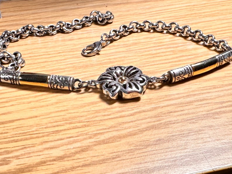 K&ouml;nnte beinhalten: Silberfarbenes Armband mit bl&uuml;tenf&ouml;rmigem Mittelst&uuml;ck und goldfarbenen Akzenten. Das Armband hat ein Kettenglied-Design und einen Karabinerverschluss. Die Blume hat eine goldfarbene Mitte.