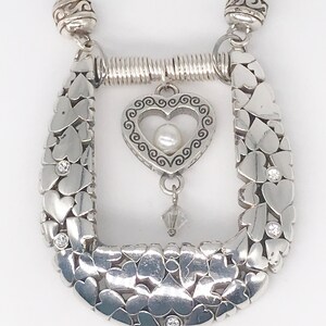 Pode incluir: Colar de prata com um pingente em forma de cora&ccedil;&atilde;o que apresenta uma p&eacute;rola e um cristal pendente. O colar &eacute; feito de uma corrente de cora&ccedil;&otilde;es entrela&ccedil;ados com pequenas pedras transparentes.