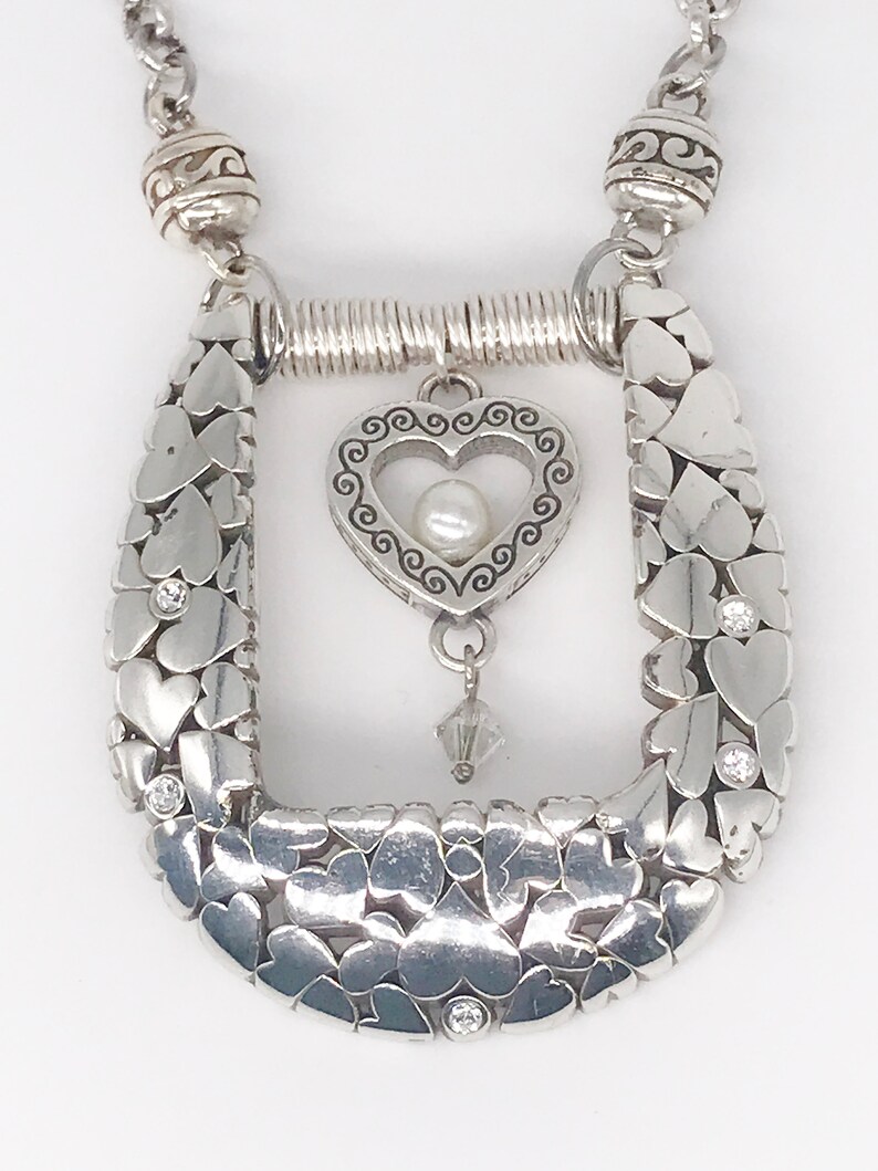 Pode incluir: Colar de prata com um pingente em forma de cora&ccedil;&atilde;o que apresenta uma p&eacute;rola e um cristal pendente. O colar &eacute; feito de uma corrente de cora&ccedil;&otilde;es entrela&ccedil;ados com pequenas pedras transparentes.