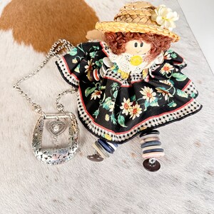 Pode incluir: Uma boneca feita &agrave; m&atilde;o vestindo um vestido floral preto e branco com um chap&eacute;u de palha e uma flor branca. A boneca est&aacute; segurando um colar de corrente de prata com um pingente em forma de cora&ccedil;&atilde;o.